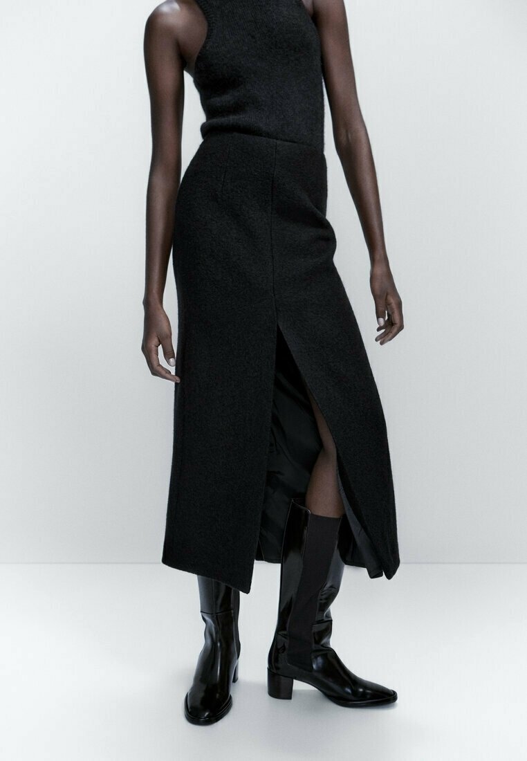 Robe midi noire sans manches avec fente frontale, portée avec des bottes à talons mi-mollet noires et brillantes, sur un mannequin debout devant un fond uni.