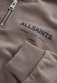 Felpa marrone con zip, con polsini a costine e una texture morbida. Presenta il marchio "ALLSAINTS" e testo giapponese. Chiusura con zip in argento.