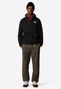 Pullover in fleece nero con zip a metà, felpa bordeaux sotto, pantaloni oliva e scarpe chiare. Presenta logo e tasca frontale.