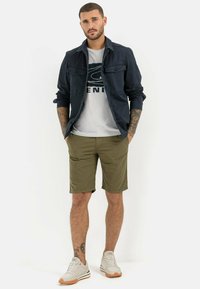 Marineblaue Hemdjacke über einem weißen Grafik-T-Shirt, olivgrüne Shorts und helle Sneakers mit orangefarbenen Akzenten; Tattoos sind am Arm sichtbar.
