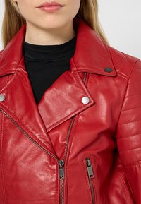 Chaqueta de motocicleta de cuero rojo con un diseño acolchado en los hombros, herrajes plateados y cierre de cremallera asimétrico. Acabado liso.