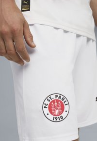 Biele futbalové šortky vyrobené z priedušného materiálu, s okrúhlym červeným a čiernym logom FC St. Pauli na ľavom stehne.