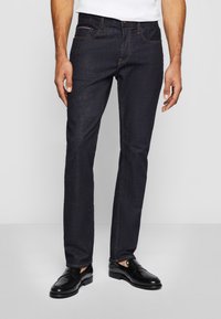 Dark blue denim jeans with a slim fit, featuring orange stitching, five pockets, and a button closure. Worn with black leather shoes.