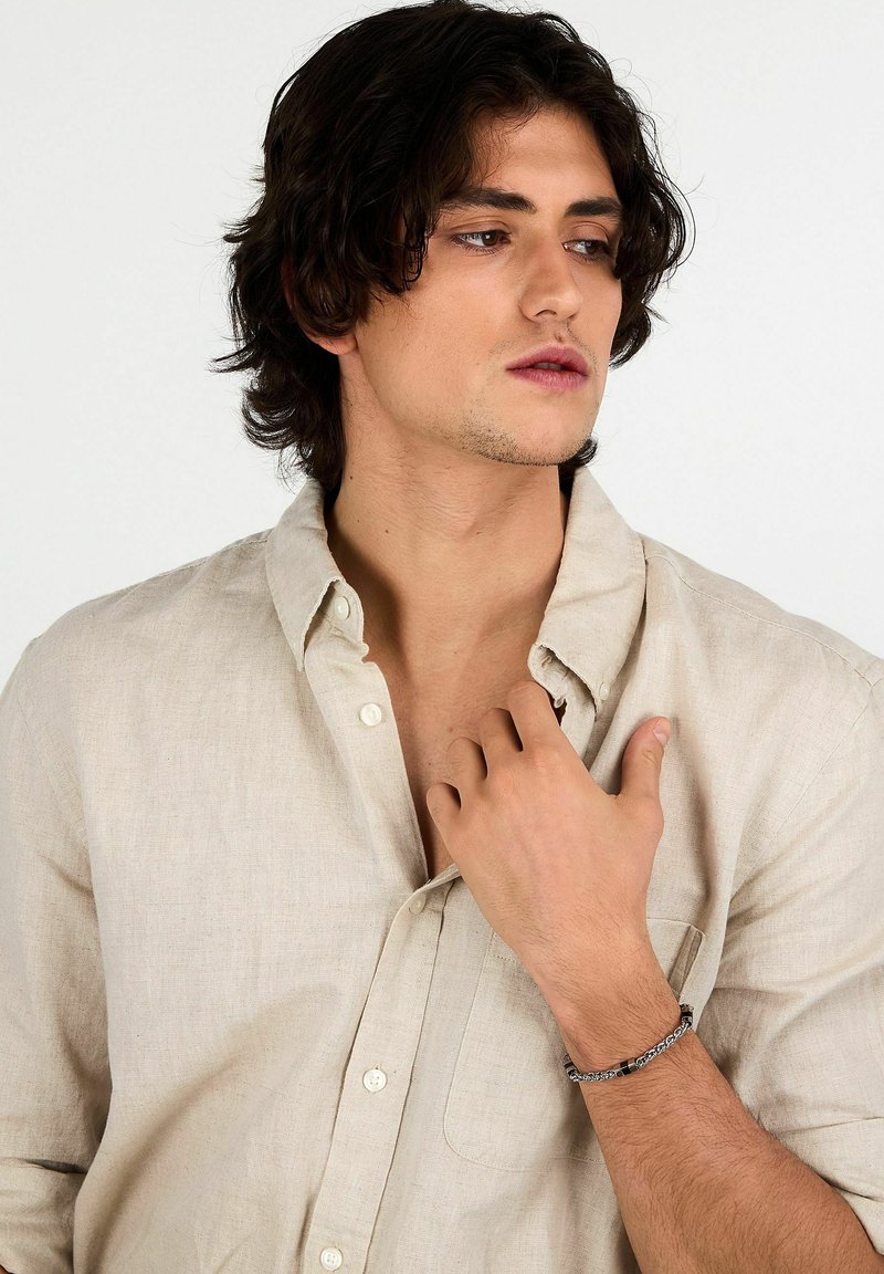 Camicia beige chiaro in lino con bottoni, colletto morbido; con braccialetto decorato al polso, pelle liscia e capelli di media lunghezza.