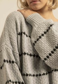Pull en tricot gris avec un tissu texturé, présentant des rayures horizontales noires et une coupe ample, accentué par des poignets et un col côtelés.