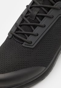 Black athletic shoe with a breathable mesh upper, textured surface, flat laces, and a smooth rubber sole; features minimalistic design elements.