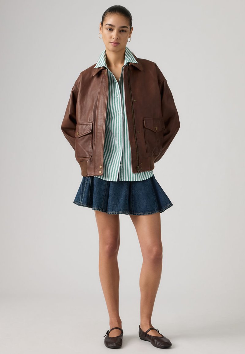 Jeune femme debout portant une veste en cuir marron, un chemisier rayé vert et blanc, une jupe en jean bleue et des chaussures Mary Jane noires.