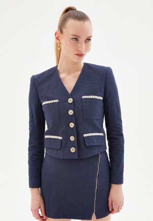 Navy cropped jacket made from textured fabric, featuring a V-neck, decorative pockets, and five gold-tone buttons with cream accents.