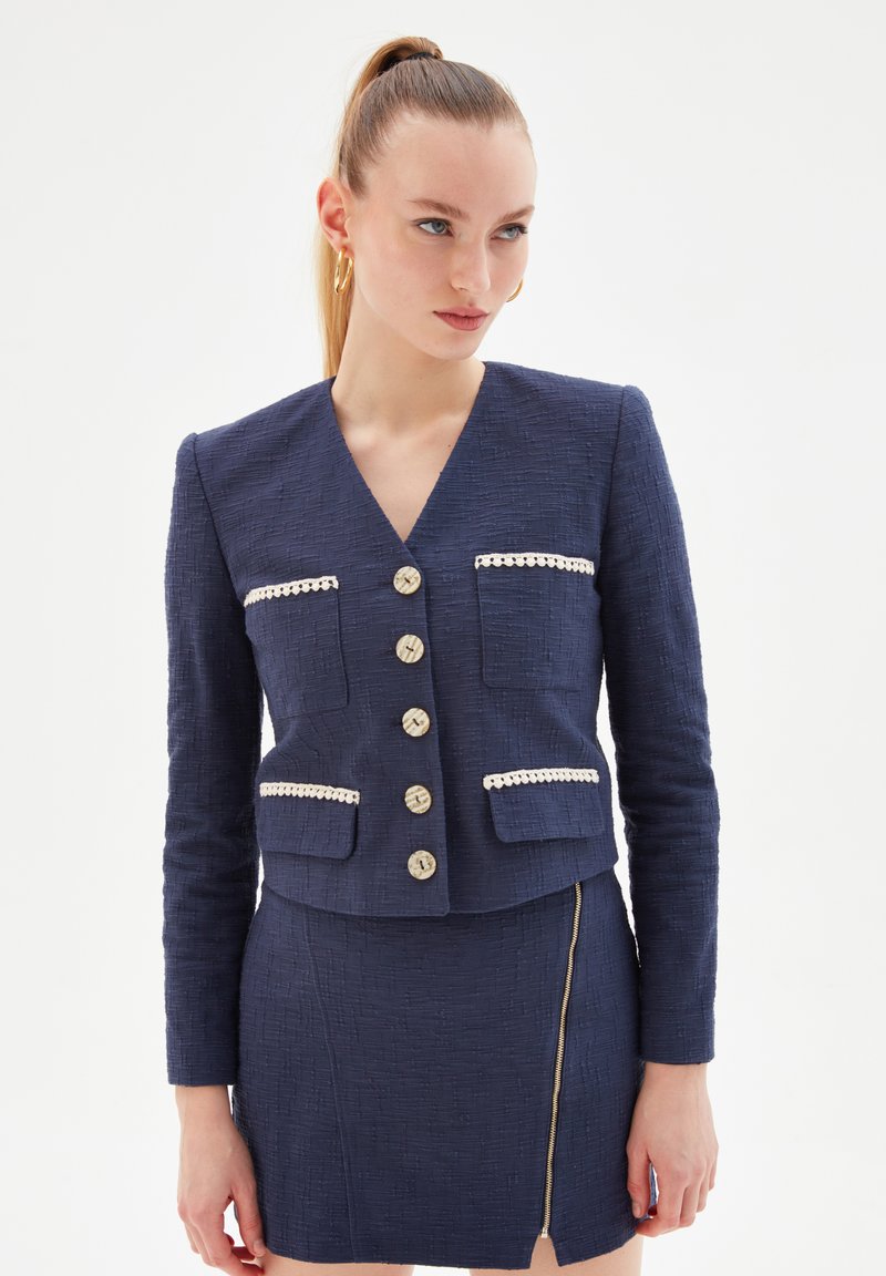 Navy cropped jacket made from textured fabric, featuring a V-neck, decorative pockets, and five gold-tone buttons with cream accents.