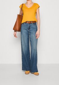 Gul v-ringad t-shirt, ljusblå vida jeans, brun bälte och brun axelväska. Modellen har på sig gula skor med en texturerad sula.