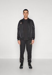 Zwart sportset bestaande uit een volledig ritssluiting jack en joggers met witte accenten. De stof lijkt glad met de "Puma" en "KING" logo's in het goud.