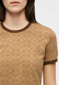 Γυναίκα που φοράει πουκάμισο κοντομάνικο με ανοιχτό καφέ σχέδιο, με σκουρότερο καφέ γιακά και περιθώρια στα μανίκια, μπροστά από ένα απλό φόντο.