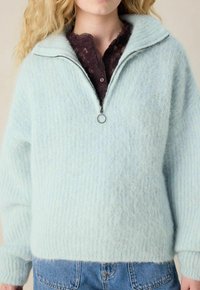 Frau in einem hellblauen, flauschigen Reißverschluss-Pullover über einem dunkelvioletten Spitzenoberteil, kombiniert mit blauen Jeans, vom Hals abwärts beschnitten.