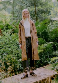 Manteau imperméable marron clair avec capuche, bord noir, porté sur un pull à motifs blancs et un pantalon gris, associé à des chaussures foncées.