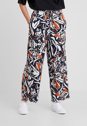 Weit geschnittene Hose mit abstrakten Mustern in Marineblau, Weiß und Rost, elastischem Bund mit Tunnelzug, getragen mit weißen Turnschuhen und einem schwarzen Oberteil.