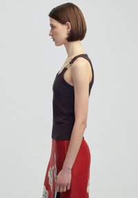 Zwarte geribbelde tanktop met een diepe halslijn, voorzien van decoratieve gouden hardware op de schouder. Gepaarde met een rode patroons rok.