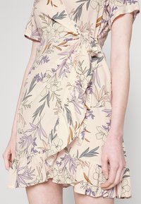 Vestido fluido color beige con patrones florales en púrpura, verde y naranja. Presenta un dobladillo con volantes y un detalle de lazo en la cintura.