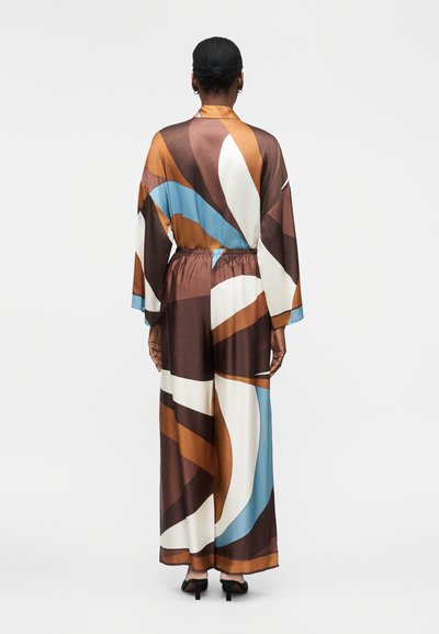 Γυναίκα με σκούρα μαλλιά σε κότσο, που φοράει ένα μακρύ, χαλαρό φόρεμα με πολύχρωμο αφηρημένο μοτίβο με καφέ, λευκά, μπλε και πορτοκαλί στοιχεία.