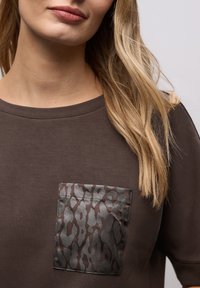 Braunes kurzärmeliges Oberteil mit einer gemusterten Tasche in dezentem Tiermuster in Grau- und Brauntönen. Glatte Textur und entspannte Passform.