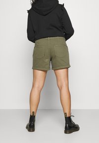 Pantalones cortos de mezclilla verde oliva con dobladillos enrollados, dos bolsillos traseros, combinados con botas negras hasta el tobillo que tienen tiradores amarillos.