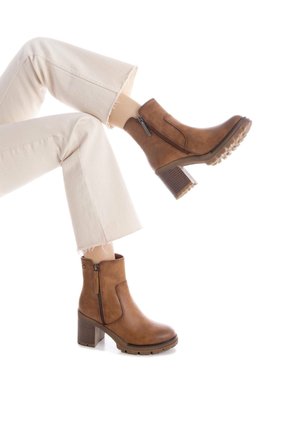 Botas de cuero marrón hasta el tobillo con cremallera lateral y suela de goma texturizada. Tacón ancho con detalles antideslizantes. Combinadas con pantalones de mezclilla de color claro.