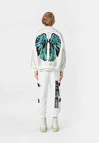 Chaqueta bomber blanca con bordado de alas en teal y negro en la parte trasera, combinada con joggers blancos con acentos negros. Botas gruesas beige.