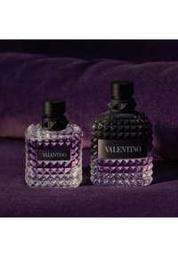 Two textured glass Valentino perfume bottles with black caps on a purple fabric surface and background.