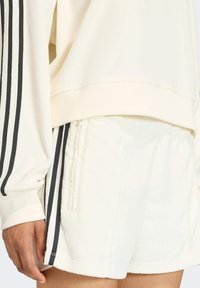 Krémová mikina so čiernymi pruhmi pozdĺž rukávov v kombinácii s krémovými šortkami so štruktúrovanou tkaninou a bočnými vreckami.