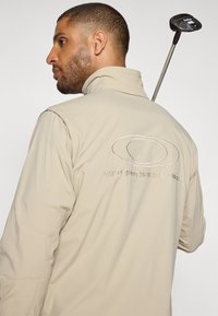 Veste légère beige avec un col haut, présentant un logo brodé et du texte au dos. Texture lisse, conçue pour le confort et la liberté de mouvement.
