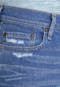 Blå denimjeans med en slitna bakficka med fransiga kanter, kopparfärgade detaljer och subtila blekningar över tyget.