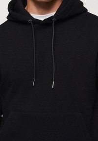 Sweat à capuche noir en tissu doux, avec une poche kangourou, des cordons terminés par des embouts métalliques, et un design simple et minimaliste.