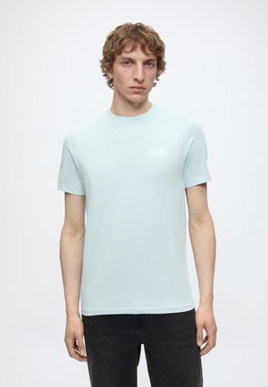Jeune homme aux cheveux bouclés châtain clair portant un t-shirt bleu pâle Karl Lagerfeld et un pantalon noir, debout devant un fond blanc uni.