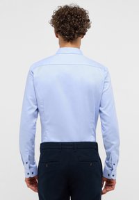Lichtblauwe overhemdd met een rechte rugpas en contrasterende donkere knopen op de manchetten, gecombineerd met donkere broek. Glad textuur van de stof.
