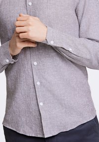 Camisa de lino gris claro con mangas largas, puños abotonados y dobladillo curvado. Presenta botones blancos y una tela texturizada.