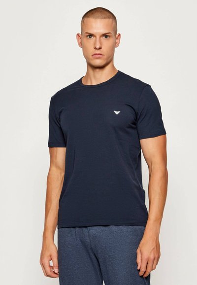 Νέος άνδρας με κοντά μαλλιά, φοράει εφαρμοστό σκούρο μπλε μπλουζάκι και μπλε παντελόνι, στέκεται μπροστά σε απλό ανοιχτό φόντο.