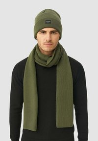 Beanie verde lavorato a maglia e sciarpa a coste abbinata, entrambi esposti su uno sfondo neutro. Il cappello presenta un'etichetta con logo; la sciarpa è lunga e strutturata.