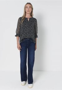 Zwarte blouse met wit abstract patroon, sleufopening en transparante mouwen. Gecombineerd met donkerblauwe flared jeans en witte sneakers.