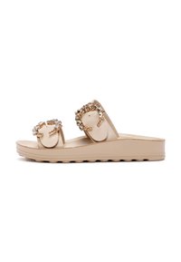 Beige slip-on sandals with two embellished straps featuring circular accents and rhinestones, on a textured sole with a slight wedge.