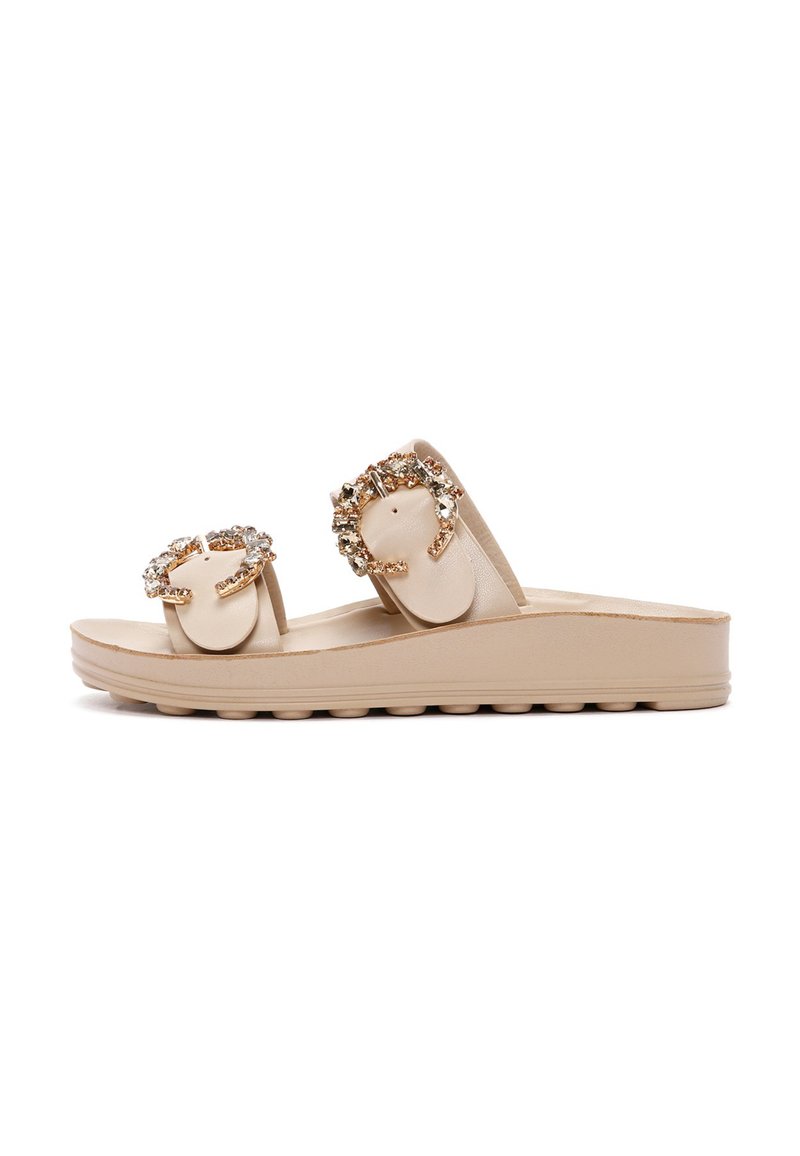 Beige slip-on sandals with two embellished straps featuring circular accents and rhinestones, on a textured sole with a slight wedge.