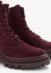 Botine din suede în nuanța burgundy, cu o talpă robustă, având un sistem de închidere cu șireturi și fermoar pe laterale pentru o purtare ușoară. Finisaj texturat și construcție solidă.