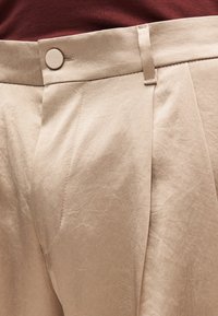 Beżowe plisowane spodnie o gładkiej fakturze, zapięcie na guzik i szlufki na pasek, szczegółowe przeszycia wzdłuż szwów.