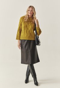 Vastag mustárszínű kötött pulóver kámzsanyakkal, térdig érő fekete bőr szoknyával és térdig érő fekete csizmával párosítva. Fekete keresztpántos táska.