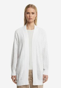 Vit stickad cardigan med öppen framsida och långa ärmar, med en strukturerad geometrisk mönstring och ribbade ärmslut. Bärs över en ljus topp.