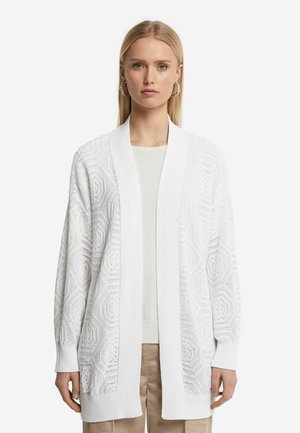 Weißer Strickcardigan mit offenem Vorderteil und langen Ärmeln, der ein strukturiertes geometrisches Muster und gerippte Bündchen aufweist. Über einem hellen Oberteil getragen.