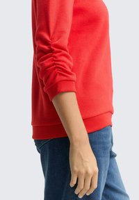 Rode gebreide sweatshirt met gatherende mouwen, ribbels aan de manchetten en zoom, gecombineerd met blauwe denim jeans. Soepele textuur, aansluitend design.