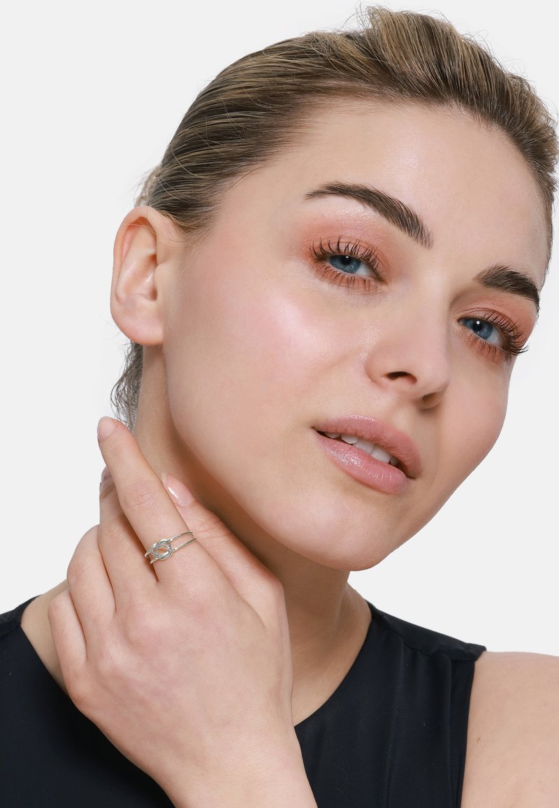 Anello d'argento con design intrecciato. Modella che lo indossa sulla mano destra, mostrando una superficie liscia e lucida. Carnagione chiara, trucco neutro.