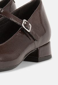 Rudos spalvos patentuotos odos Mary Jane stiliaus bateliai su žemu blokiniu kulnu, vienu dirželiu ir sidabrine sagties detalė. Lygi tekstūra, apvalus priekis.