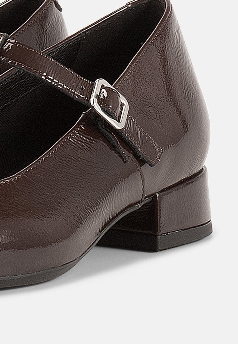 Brown patent leather Mary Jane shoe with a low block heel, featuring a single strap and silver buckle detail. Smooth texture, rounded toe.
