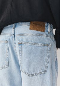 Lichtblauwe denim jeans met achterzakken en een bruin leren label met de tekst "PULL&BEAR", gedragen met een zwart shirt.