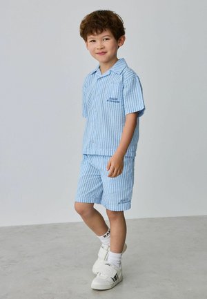 Junger Junge trägt ein blau gestreiftes kurzärmeliges Hemd und passende Shorts, weiße Socken und weiße Turnschuhe, steht auf einem grauen Boden.